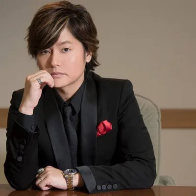 Showtaro Morikubo สวมสูทสีดำพร้อมผ้าเช็ดหน้าสีแดง นั่งเท้าคางอยู่ที่โต๊ะไม้